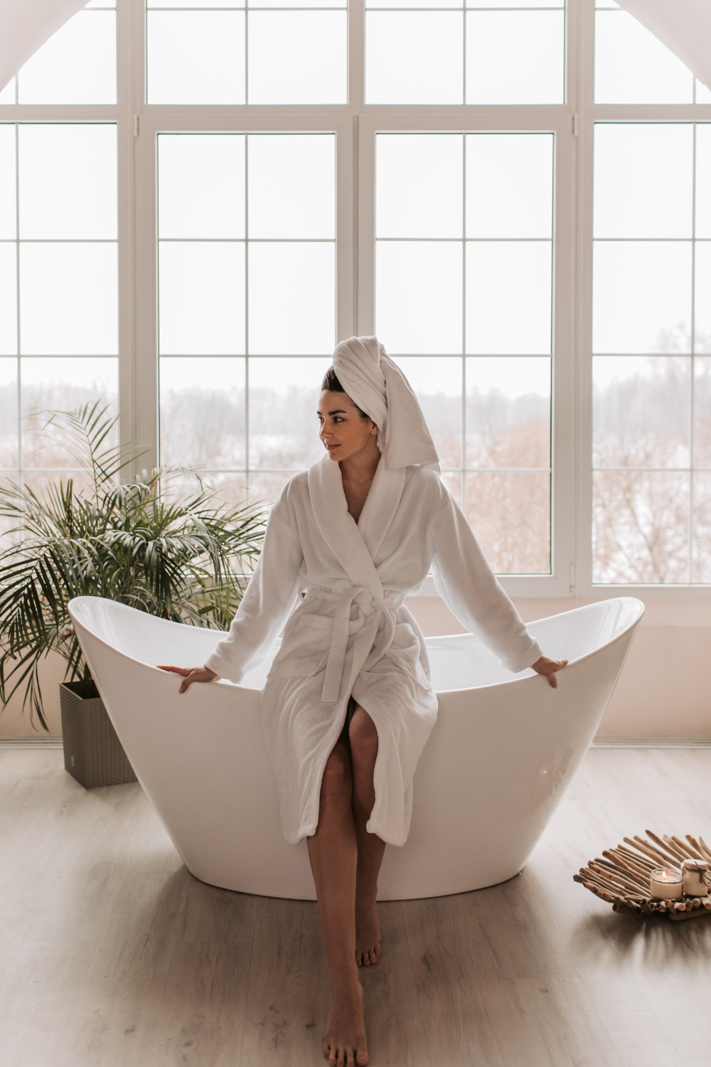 Woman in a white bathrobe sitting on the edge of a modern bathtub in a serene spa setting.